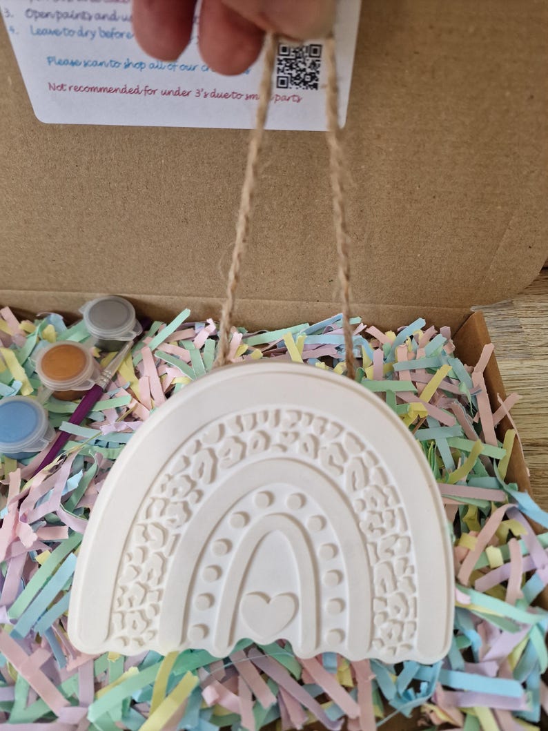 May include: A white ceramic rainbow ornament with a heart and leopard print design, hanging by a twine string. The ornament is in a cardboard box filled with colorful paper shreds, with paint pots and a brush visible.