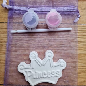 May include: A purple mesh bag with a white ceramic crown that says "Princess" on it. Two small containers of paint, one purple and one pink, are included.