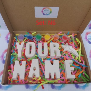 May include: A craft kit in a cardboard box, featuring the words "YOUR NAME" in large, unpainted letters. The kit includes small paint pots in various colors, a star-shaped figure, and colorful paper shreds for decoration. The box is labeled "Set: N6".