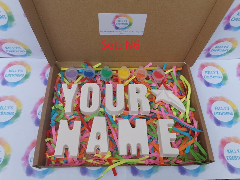 May include: A craft kit in a cardboard box, featuring the words "YOUR NAME" in large, unpainted letters. The kit includes small paint pots in various colors, a star-shaped figure, and colorful paper shreds for decoration. The box is labeled "Set: N6".