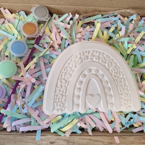 May include: A craft kit featuring a white, textured rainbow with a heart design, paint pots in various colors, and a paintbrush. The kit is nestled in a cardboard box filled with pastel-colored paper shreds.
