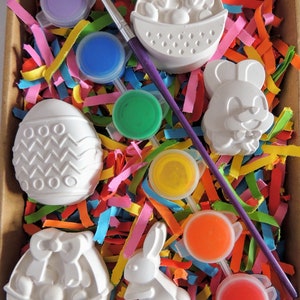 May include: A set of six white ceramic Easter-themed figurines, including a bunny, an egg, and a basket, with six paint pots in various colors, including purple, blue, green, yellow, orange, and red, and a paintbrush.