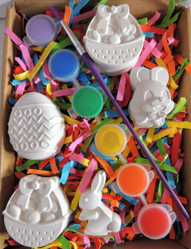 May include: A set of six white ceramic Easter-themed figurines, including a bunny, an egg, and a basket, with six paint pots in various colors, including purple, blue, green, yellow, orange, and red, and a paintbrush.