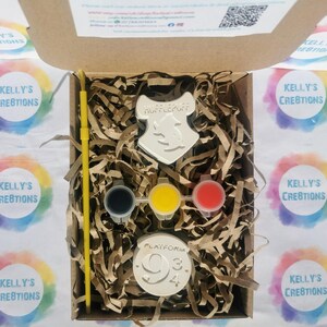 May include: A craft kit in a cardboard box, featuring a Hufflepuff crest, Platform 9 3/4 emblem, paint pots, and a yellow paintbrush. The kit includes brown paper filler. The box is surrounded by colorful stickers.