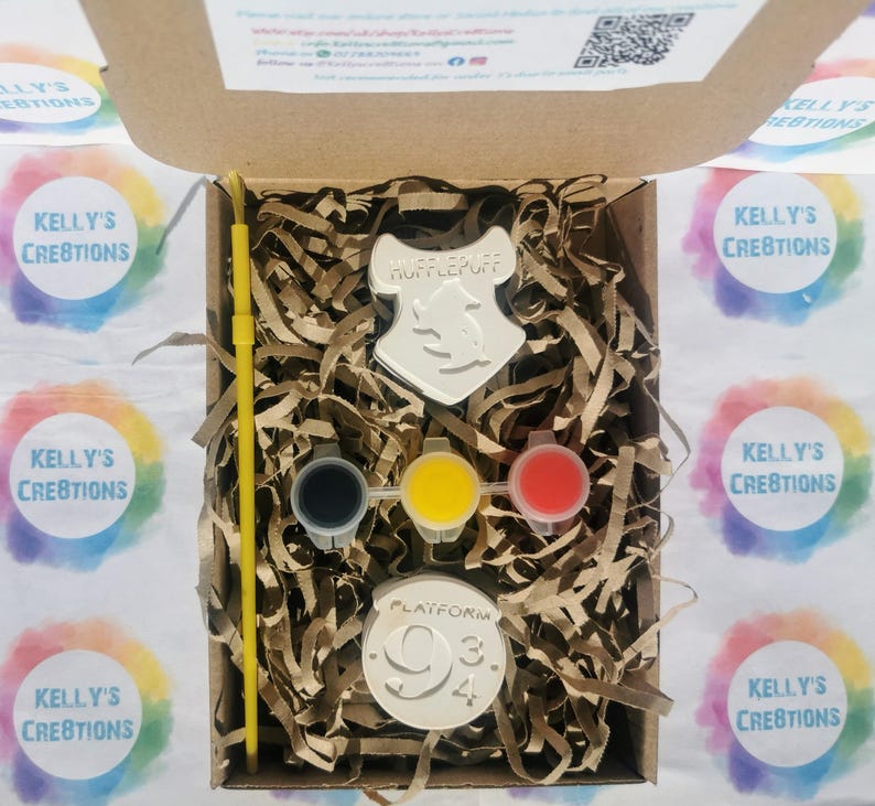 May include: A craft kit in a cardboard box, featuring a Hufflepuff crest, Platform 9 3/4 emblem, paint pots, and a yellow paintbrush. The kit includes brown paper filler. The box is surrounded by colorful stickers.