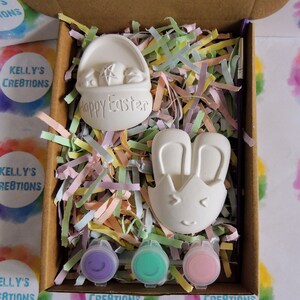 May include: A craft kit containing two white ceramic Easter-themed figurines, a basket and a bunny, and three small paint pots in purple, green, and pink. The kit is packaged in a brown box with colourful shredded paper.