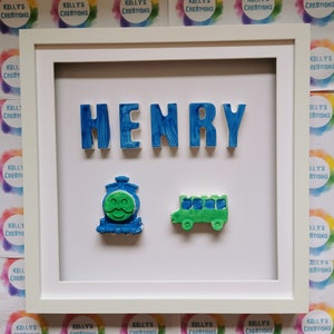 May include: A framed piece of art displaying the name "HENRY" in blue, textured letters. Beneath the name are a blue train with a green face and a green bus. The frame is white, with "KELLY'S CREATIONS" in the background.