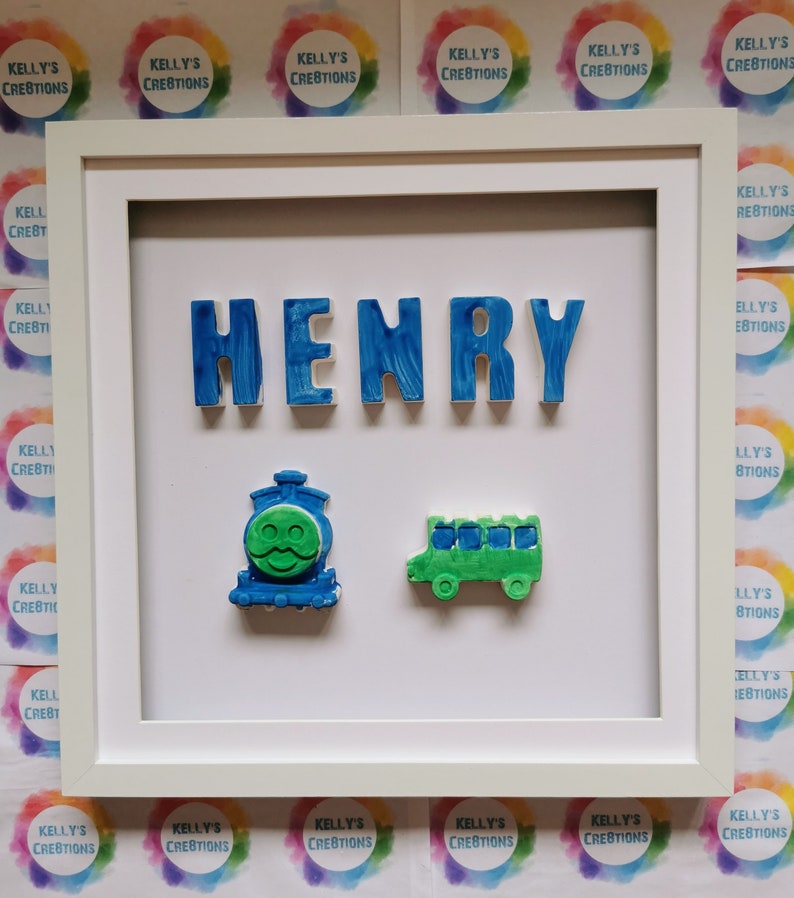 May include: A framed piece of art displaying the name "HENRY" in blue, textured letters. Beneath the name are a blue train with a green face and a green bus. The frame is white, with "KELLY'S CREATIONS" in the background.