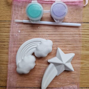 Puede incluir: Un conjunto de dos botes de pintura, uno azul verdoso y otro morado, con un pincel y tres formas de yeso blanco: un arco&iacute;ris, una nube y una estrella fugaz. Los art&iacute;culos est&aacute;n en una bolsa de malla rosa.