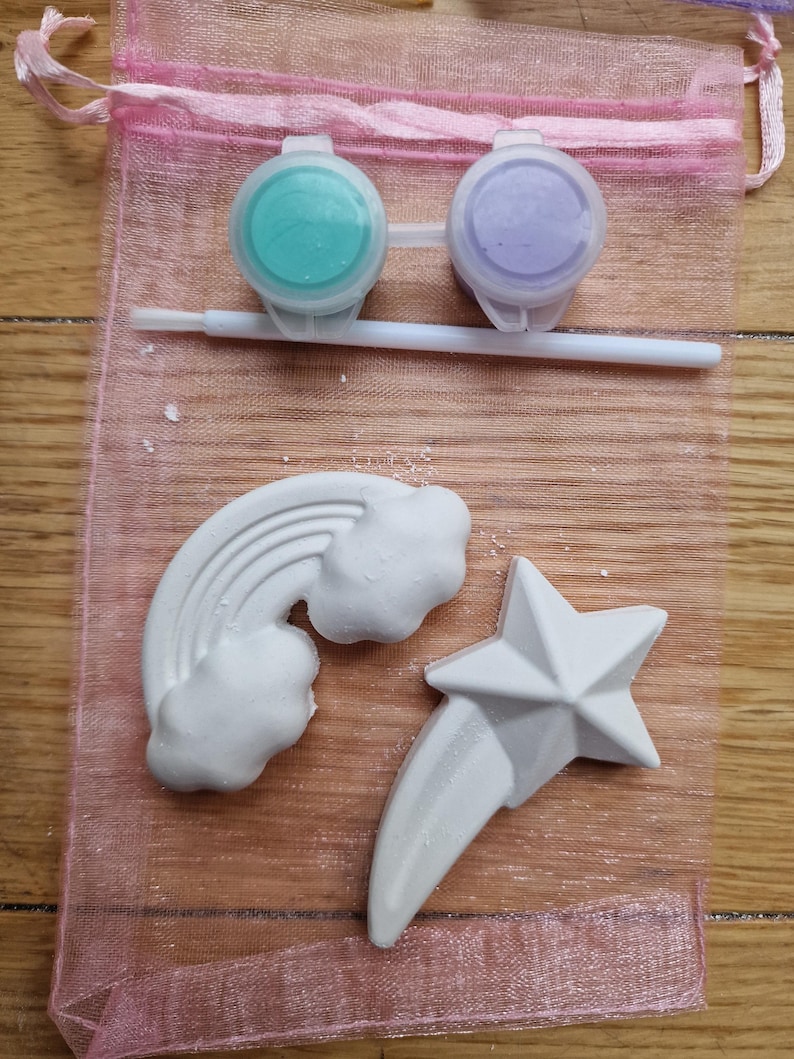 Puede incluir: Un conjunto de dos botes de pintura, uno azul verdoso y otro morado, con un pincel y tres formas de yeso blanco: un arco&iacute;ris, una nube y una estrella fugaz. Los art&iacute;culos est&aacute;n en una bolsa de malla rosa.