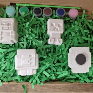 Puede incluir: Un kit de manualidades con figuras de bloques blancos, botes de pintura de varios colores y un pincel verde. Las figuras est&aacute;n dispuestas sobre tiras de papel verde dentro de una caja de cart&oacute;n.