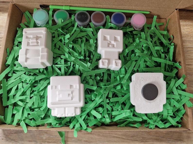 Puede incluir: Un kit de manualidades con figuras de bloques blancos, botes de pintura de varios colores y un pincel verde. Las figuras est&aacute;n dispuestas sobre tiras de papel verde dentro de una caja de cart&oacute;n.