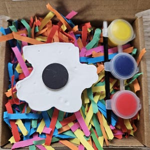 May include: A craft kit in a cardboard box, featuring a white cloud-shaped magnet, three small paint pots with yellow, blue, and red paint, and a paintbrush. The box is filled with colourful shredded paper. Text on the box reads: "Not recommended for under 3's due to small parts."