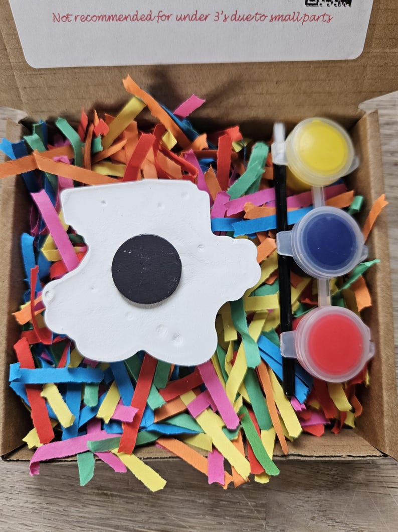 May include: A craft kit in a cardboard box, featuring a white cloud-shaped magnet, three small paint pots with yellow, blue, and red paint, and a paintbrush. The box is filled with colourful shredded paper. Text on the box reads: "Not recommended for under 3's due to small parts."