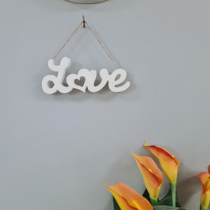 May include: White wooden sign with the word "Love" in cursive with a heart in the middle of the word. The sign is hanging from a string on a grey wall. There are orange calla lilies in the bottom right corner of the image.