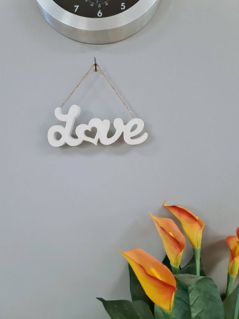 May include: White wooden sign with the word "Love" in cursive with a heart in the middle of the word. The sign is hanging from a string on a grey wall. There are orange calla lilies in the bottom right corner of the image.