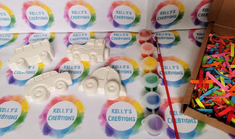 May include: A craft kit with four white ceramic cars, a set of six paint pots, a paintbrush, and colorful shredded paper in a box. The cars are ready to be painted.