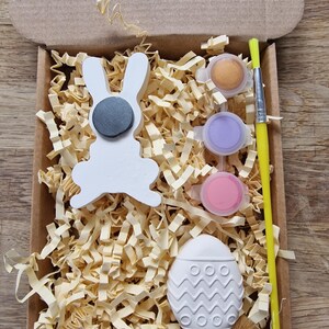 May include: A craft kit for painting a white ceramic bunny and egg. The kit includes three small pots of paint in gold, purple, and pink, a paintbrush, and a small box filled with shredded paper.