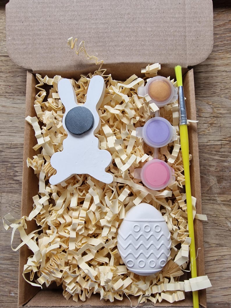 May include: A craft kit for painting a white ceramic bunny and egg. The kit includes three small pots of paint in gold, purple, and pink, a paintbrush, and a small box filled with shredded paper.