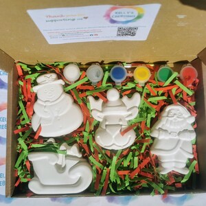 May include: A craft kit in a cardboard box, featuring unpainted ceramic Christmas ornaments: a snowman, reindeer, Santa, and a sleigh. The kit includes paint pots in various colors and decorative paper. The box has the text "Kelly's Creations".