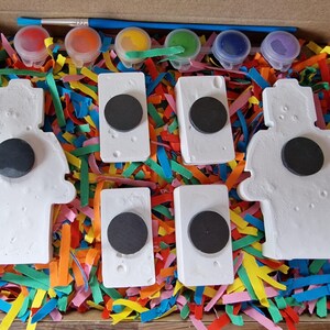 May include: A set of five white ceramic figurines with black circles in the center, ready to be painted. The figurines are surrounded by colorful shredded paper and five small bottles of paint.
