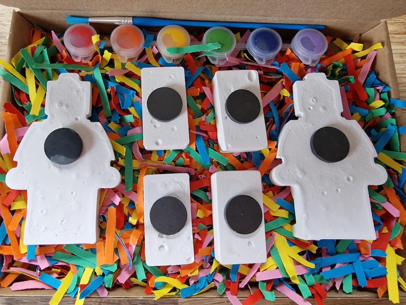 May include: A set of five white ceramic figurines with black circles in the center, ready to be painted. The figurines are surrounded by colorful shredded paper and five small bottles of paint.