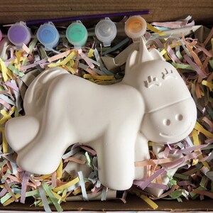 May include: A white ceramic unicorn figurine in a cardboard box with colorful shredded paper. The unicorn has a smiling face and closed eyes. Five small paint pots with pink, purple, blue, green, and orange paint are included.