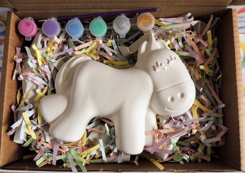 May include: A white ceramic unicorn figurine in a cardboard box with colorful shredded paper. The unicorn has a smiling face and closed eyes. Five small paint pots with pink, purple, blue, green, and orange paint are included.