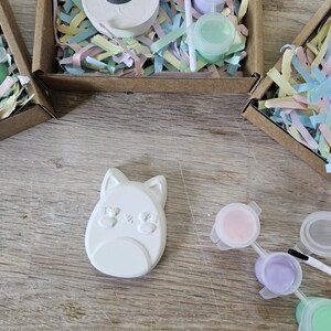 Puede incluir: Un im&aacute;n de yeso blanco con forma de gato y acabado liso, acompa&ntilde;ado de un pincel peque&ntilde;o y tres botes de pl&aacute;stico con pintura rosa, morada y verde, todo dentro de una caja de cart&oacute;n marr&oacute;n con relleno de papel de colores.