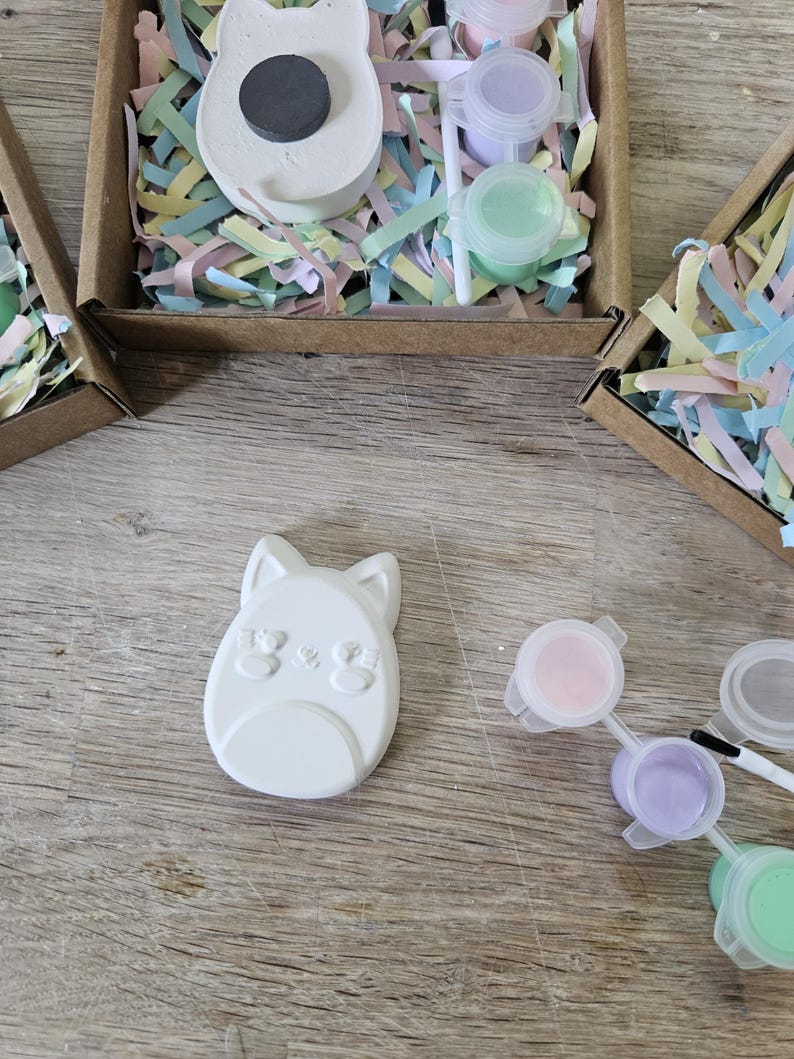 Puede incluir: Un im&aacute;n de yeso blanco con forma de gato y acabado liso, acompa&ntilde;ado de un pincel peque&ntilde;o y tres botes de pl&aacute;stico con pintura rosa, morada y verde, todo dentro de una caja de cart&oacute;n marr&oacute;n con relleno de papel de colores.