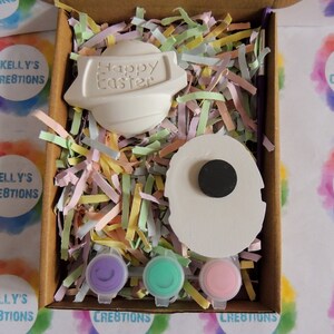 May include: A craft kit containing two white ceramic Easter eggs, one with the text "Happy Easter" and the other with a black circle. The kit includes three small pots of paint in purple, green, and pink.