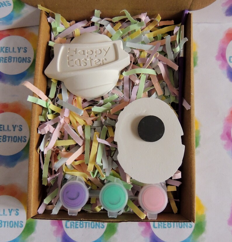 May include: A craft kit containing two white ceramic Easter eggs, one with the text "Happy Easter" and the other with a black circle. The kit includes three small pots of paint in purple, green, and pink.