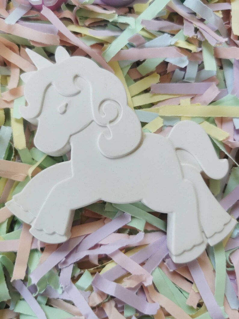 May include: A white, three-dimensional unicorn figurine with a horn, mane, and tail. The unicorn is set against a background of pastel-colored paper strips. The figurine is a blank canvas, ready for painting or decorating.