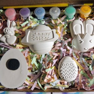 May include: A set of five ceramic Easter-themed figurines, including a bunny, an egg, and a sign that reads "Happy Easter". The figurines are white and are surrounded by colorful shredded paper. Five small bottles of paint are included.