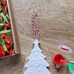 May include: White ceramic Christmas tree ornament with a star on top and red and white twine for hanging. The ornament is ready to be painted with red and green paint.