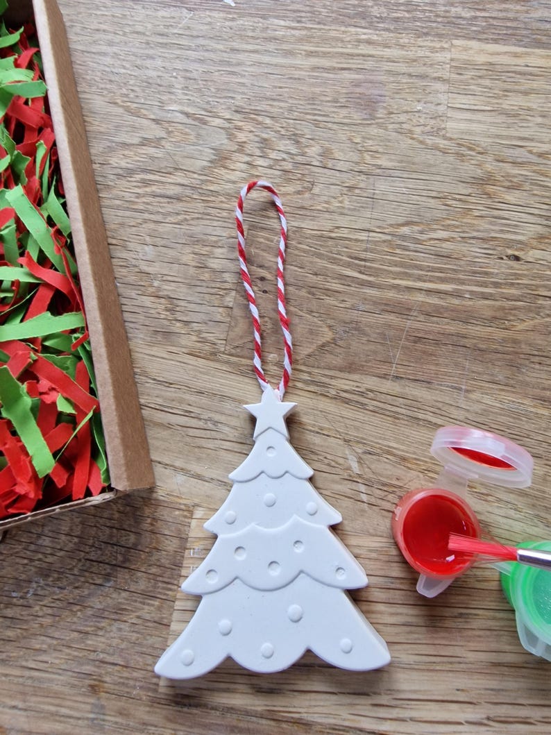 May include: White ceramic Christmas tree ornament with a star on top and red and white twine for hanging. The ornament is ready to be painted with red and green paint.