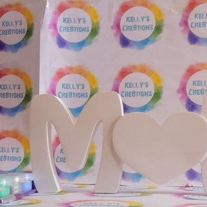 Pu&ograve; includere: Un kit per lavoretti per dipingere un cartello in legno "MAMMA" con un cuore. Il kit include barattoli di vernice di vari colori e una scatola di cartone con un'etichetta che dice "Kelly's Creations".
