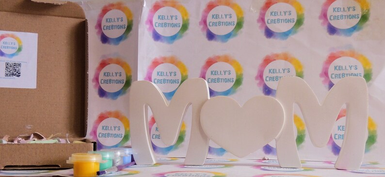Pu&ograve; includere: Un kit per lavoretti per dipingere un cartello in legno "MAMMA" con un cuore. Il kit include barattoli di vernice di vari colori e una scatola di cartone con un'etichetta che dice "Kelly's Creations".