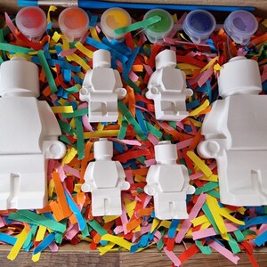 May include: A set of five white ceramic figurines shaped like Lego people, with paint pots and brushes for painting. The figurines are surrounded by colorful paper shreds.
