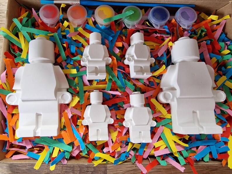 May include: A set of five white ceramic figurines shaped like Lego people, with paint pots and brushes for painting. The figurines are surrounded by colorful paper shreds.
