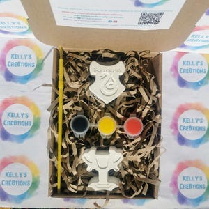 May include: A craft kit in a cardboard box, featuring a Slytherin crest and a trophy-shaped item. The kit includes paint pots in black, yellow, and red, along with a yellow paintbrush. The box is filled with shredded paper.