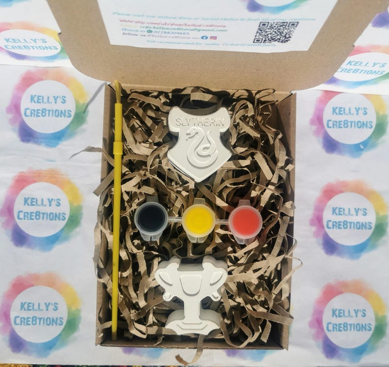 May include: A craft kit in a cardboard box, featuring a Slytherin crest and a trophy-shaped item. The kit includes paint pots in black, yellow, and red, along with a yellow paintbrush. The box is filled with shredded paper.