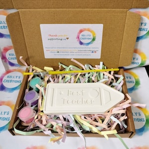 May include: A white ceramic pencil-shaped plaque with the text "Best Teacher" in black lettering, paint pots, a paintbrush, and colorful shredded paper in a cardboard box.
