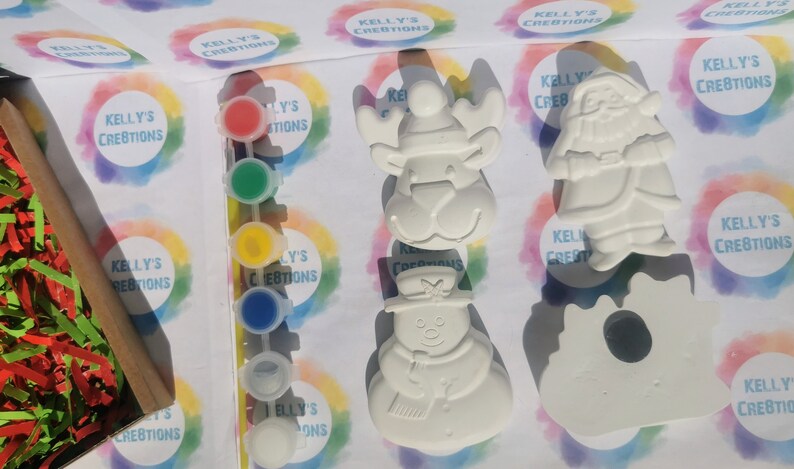 May include: A paint-your-own Christmas craft kit featuring a reindeer, snowman, and Santa Claus figurine. The kit includes a set of paint pots in various colors and the text "KELLY'S CREBTIONS" on the packaging.