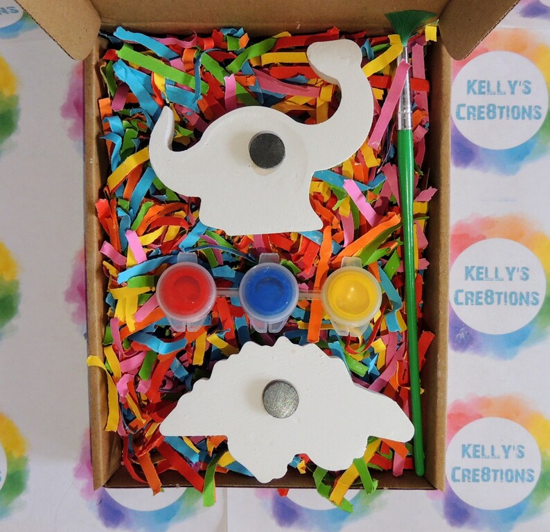 Op de afbeelding: Een knutselpakket met dinosaurus thema in een kartonnen doos. Het bevat twee witte dinosaurusvormen met magneten, drie verfpotjes (rood, blauw, geel) en een groene verfkwast. De doos is gevuld met kleurrijk papier. De tekst "KELLY'S CREATIONS" is zichtbaar.