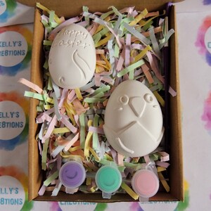 May include: A craft kit containing two white ceramic eggs, one with the words "Happy Easter" and the other with a bird design. The eggs are in a box filled with colourful shredded paper and three small pots of paint: purple, green, and pink.