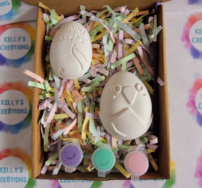 May include: A craft kit containing two white ceramic eggs, one with the words "Happy Easter" and the other with a bird design. The eggs are in a box filled with colourful shredded paper and three small pots of paint: purple, green, and pink.