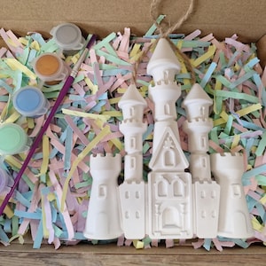 May include: A white ceramic castle kit with paint pots and a paintbrush, nestled in a cardboard box filled with colorful paper strips. The paint colors include pink, purple, green, blue, orange, and gray.