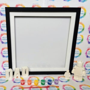May include: A paint-by-numbers kit featuring a white LEGO figure and the word "DAD" in white letters. The kit includes a black frame, six small paint pots, and a paintbrush.