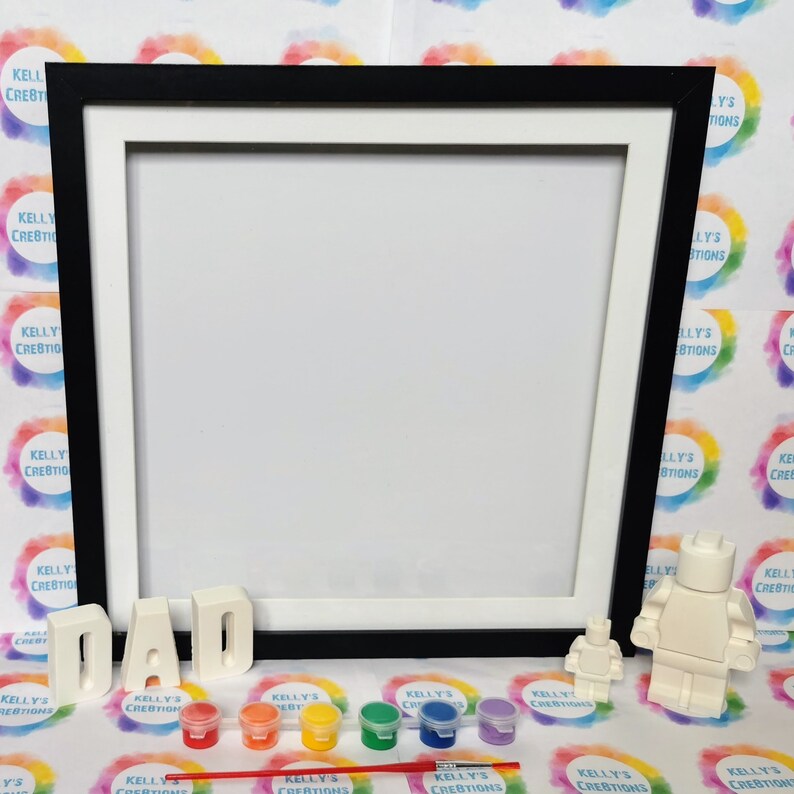 May include: A paint-by-numbers kit featuring a white LEGO figure and the word "DAD" in white letters. The kit includes a black frame, six small paint pots, and a paintbrush.